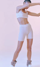 Load image into Gallery viewer, Woman standing wearing activewear with NAGNATA Balati Biker short in cream sandstone colourway, available at Amara Home.
