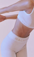 Load image into Gallery viewer, Woman standing wearing activewear with NAGNATA Balati Biker short in cream sandstone colourway, available at Amara Home.
