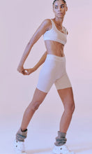 Load image into Gallery viewer, Woman standing wearing activewear with NAGNATA Balati Biker short in cream sandstone colourway, available at Amara Home.
